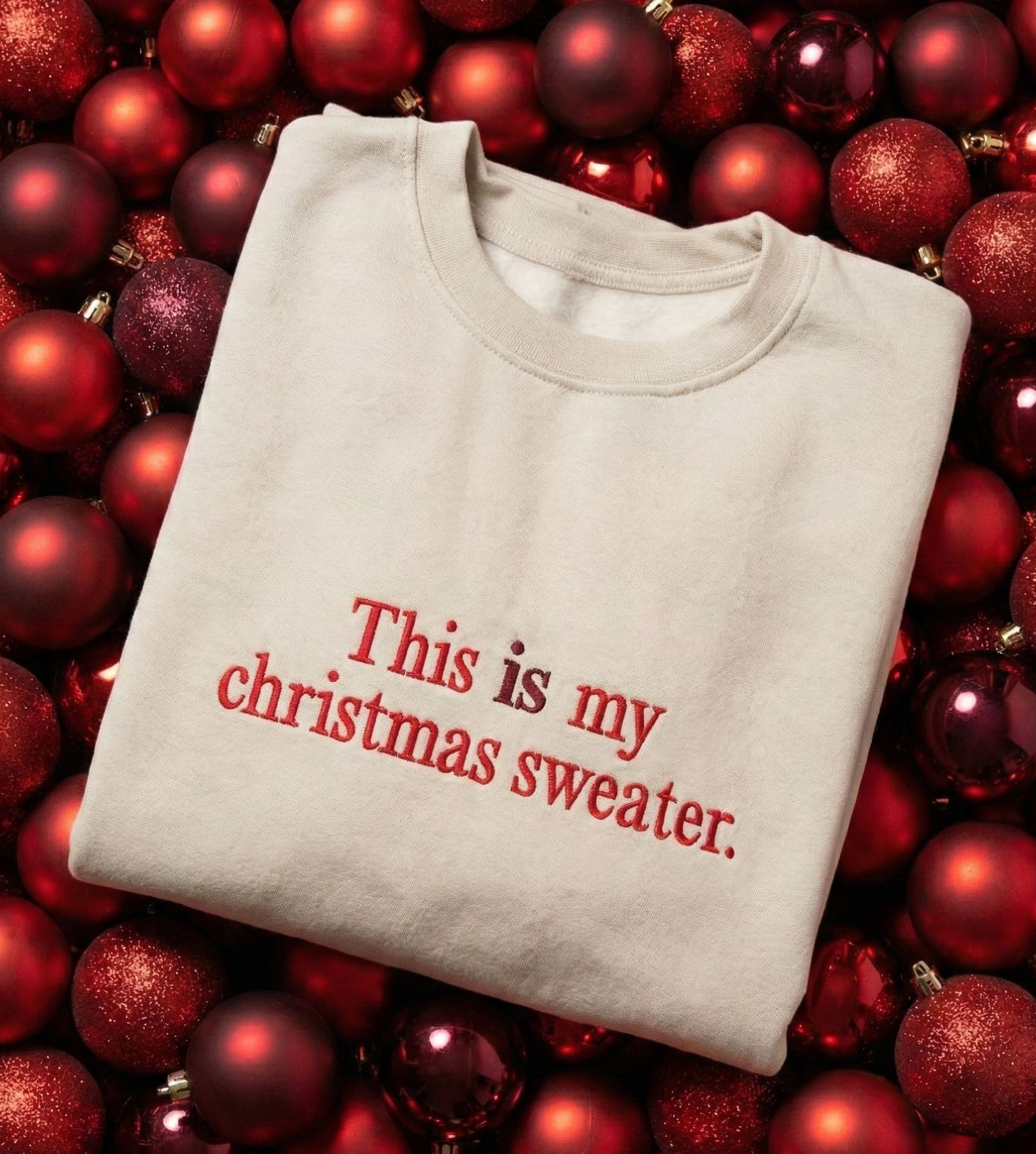 Folded beige sweater with 'This is my Christmas sweater' text on red Christmas ornaments.